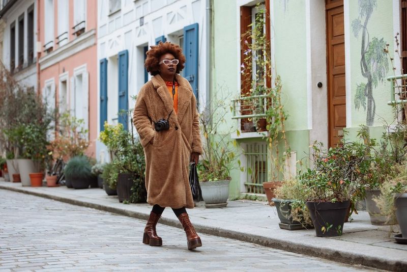 L'influencer Fatou N'Diaye, con un soffice cappotto marrone e un paio di occhiali, passa davanti a delle case color pastello portando con sé Canon EOS R50.
