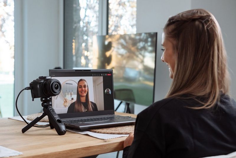 A woman filming herself with a Canon EOS R50 on a tripod grip. The footage can be seen streaming on the screen of a laptop on the desk in front of her.