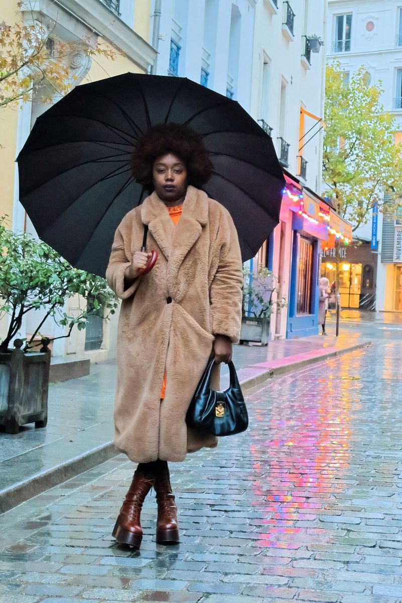 L'influencer Fatou N'Diaye con un soffice cappotto marrone e un ombrello, in piedi su una strada accanto alle vetrine illuminate.