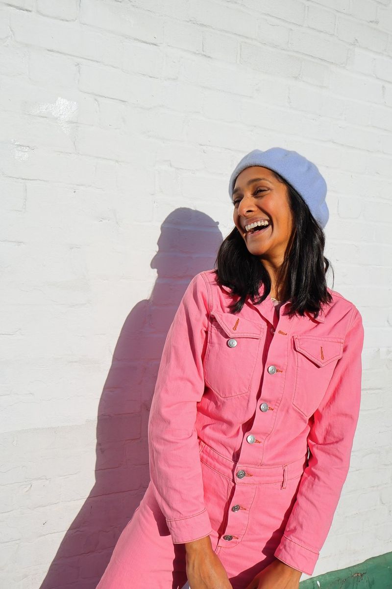 A portrait taken on a Canon EOS R50 of style influencer Zeena Shah wearing a pink jumpsuit and blue hat. She is standing against a white-painted wall and laughing.
