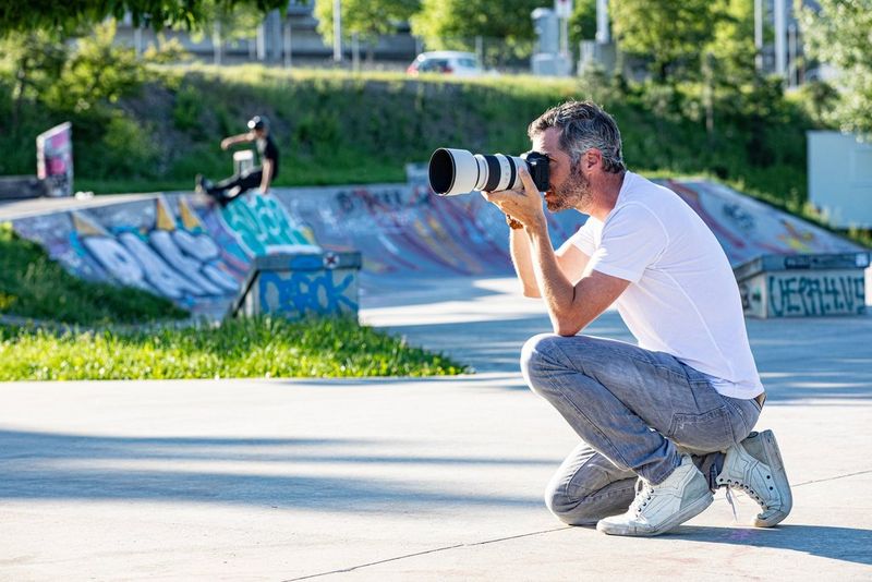 A photographer on one knee in a skate park, holding a Canon EOS R5 with a zoom lens to his eye.