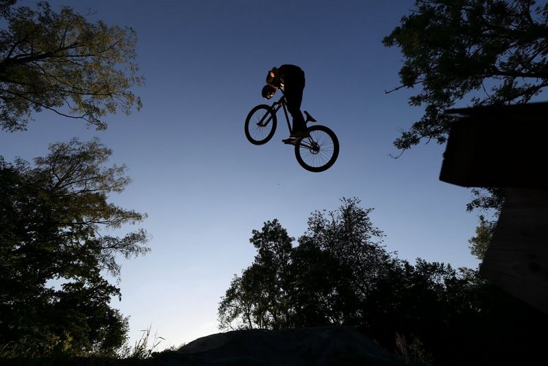 A cyclist leaps overhead, framed by trees that are silhouetted against a blue sky.