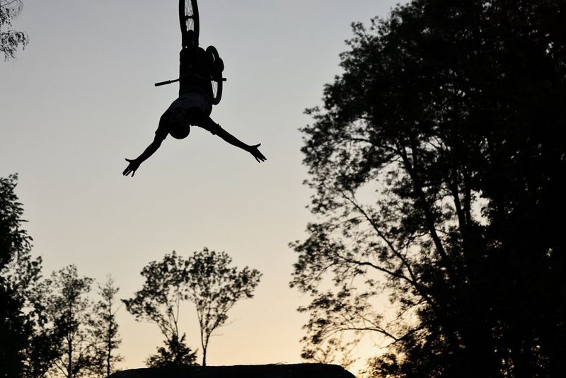 A mountain biker mid-stunt and upside down, silhouetted against the sky and framed by trees.