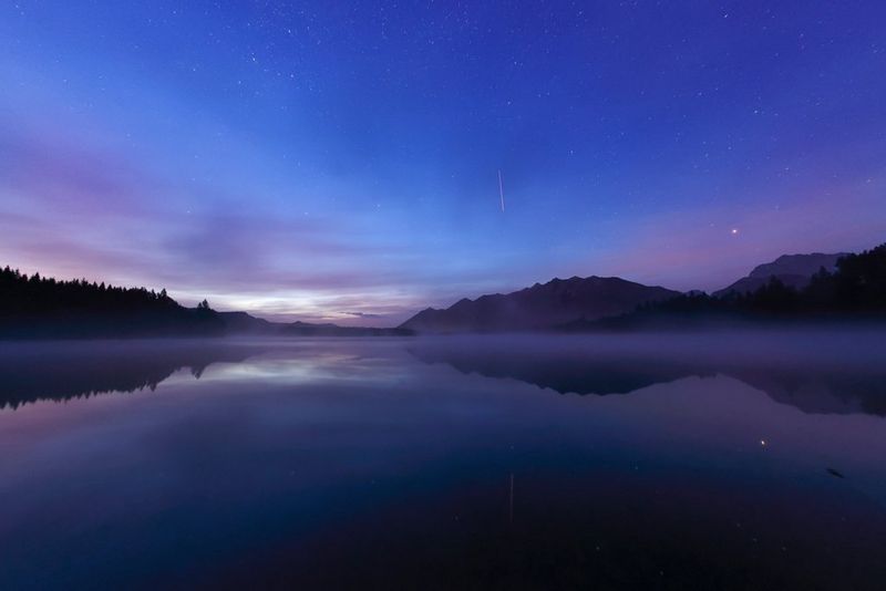 Ночное небо над озером, на поверхности которого отражаются окружающие его холмы и деревья. В небе можно увидеть звезды и метеоры.