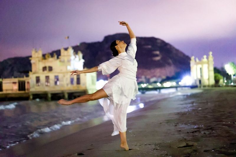 A photo taken on a Canon EOS R6 camera, showing a woman in a white dress dancing on a beach at night with buildings lit up in the background.