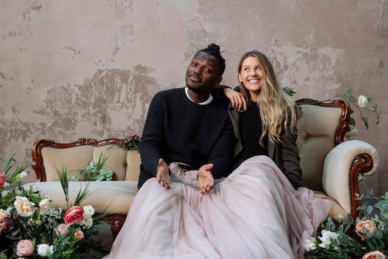 Presenter and content creator Tomi Adebayo and photographer Rosie Hardy sit together on a chaise longue, a light pink ballgown arranged over their knees and flowers on either side.