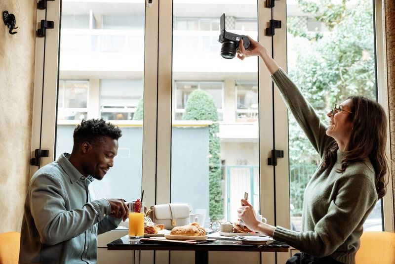 Social media content creator Laura Hannoun extends an arm holding a Canon EOS R6 Mark II pointing down at food on a table while a man sitting across the table eats his food. 