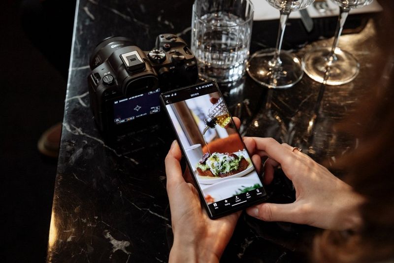 A pair of hands holds a smartphone showing the Canon Camera Connect app displaying a photo of food taken on a Canon EOS R6 Mark II