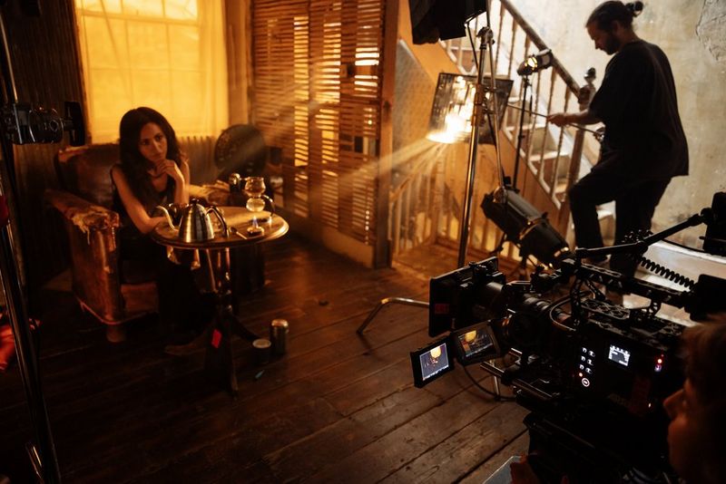 Persona seduta in poltrona, con la luce che filtra dalle persiane conferendo alla scena una sfumatura arancione, mentre la telecamera e l'obiettivo Canon riprendono da destra.