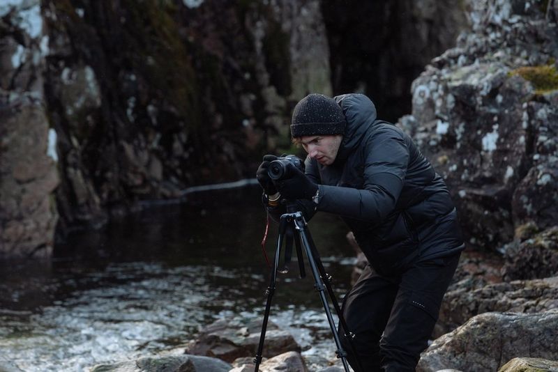 A photographer in winter clothing crouches behind a Canon EOS R6 mounted on a tripod positioned on rocks alongside a river. 