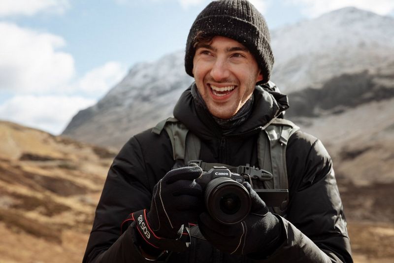 Scatto di Harrison Brown con montagne sullo sfondo che tiene in mano una fotocamera Canon e sorride.