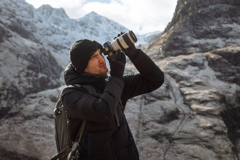 Harrison Brown, lo sguardo rivolto verso il suo soggetto in alto, guarda nel mirino di una fotocamera Canon mentre alle sue spalle si stagliano delle montagne rocciose.