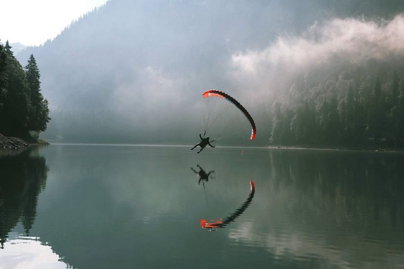 A paraglider flies low over a calm mountain lake, reflected in the water as mist hangs over the surrounding forested slopes.
