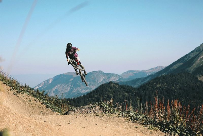 A mountain biker jumps into the air above a dirt trail, with forested mountains and a clear blue sky in the background.