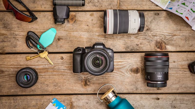 Photo en plongée d'un Canon EOS R6 Mark II posé sur une surface en bois, entouré d'objectifs Canon, de lunettes de soleil, d'une carte, d'un trousseau de clés et d'une bouteille d'eau.