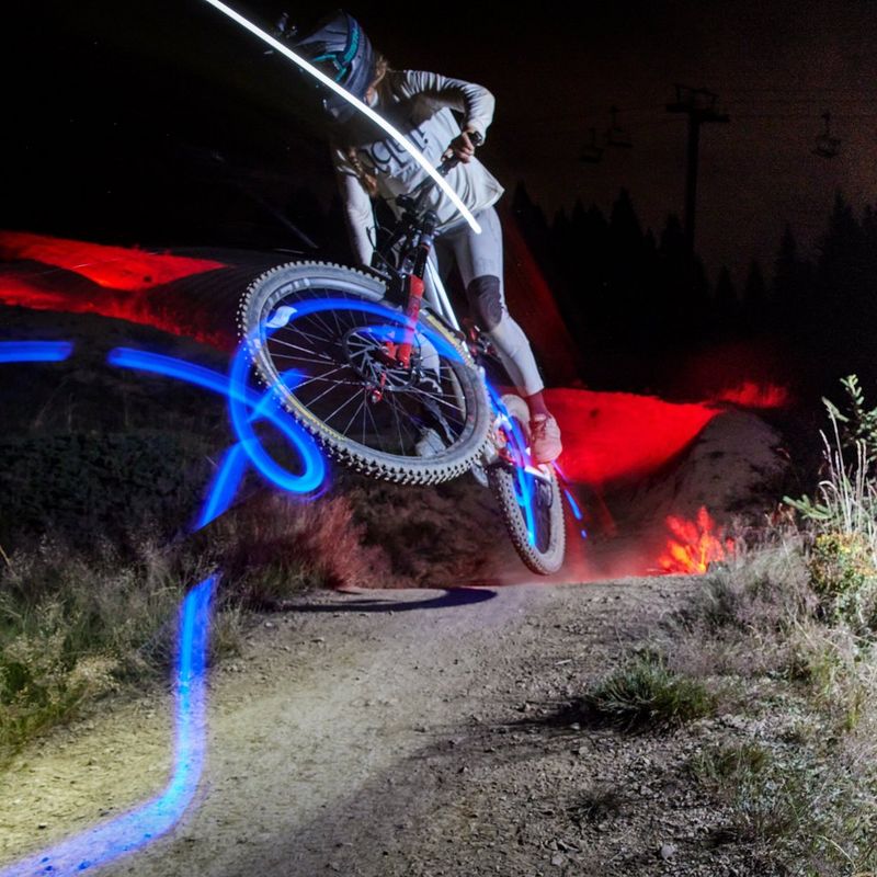 A photographer with a camera on a tripod photographs a mountain bike rider performing a stunt in mid-air at night.