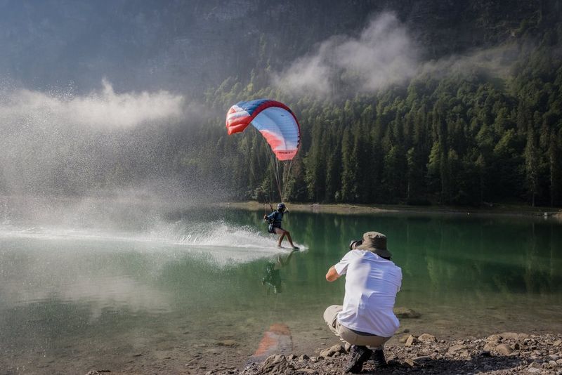 Le photographe sportif Teddy Morellec accroupi au bord d'un lac avec son Canon EOS R6 Mark II pour photographier un speed flyer frôlant la surface de l'eau.