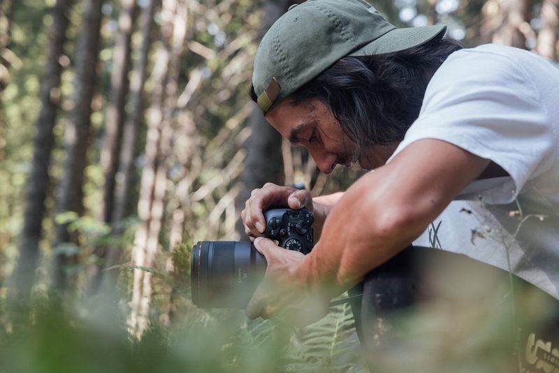 A man crouches down in grass, filming on a Canon EOS R6 Mark II.