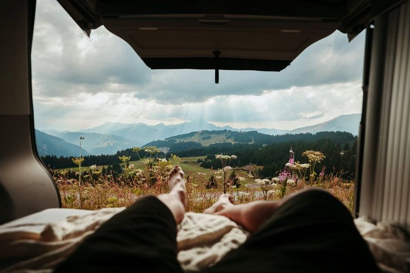 A person's legs outstretched in the back of a van with the car boot open, looking over a scenic mountain view.