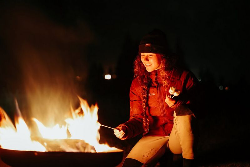 A woman in winter clothing smiles as she roasts a marshmallow on a stick over a fire at night. Taken on a Canon EOS R6 Mark II. 