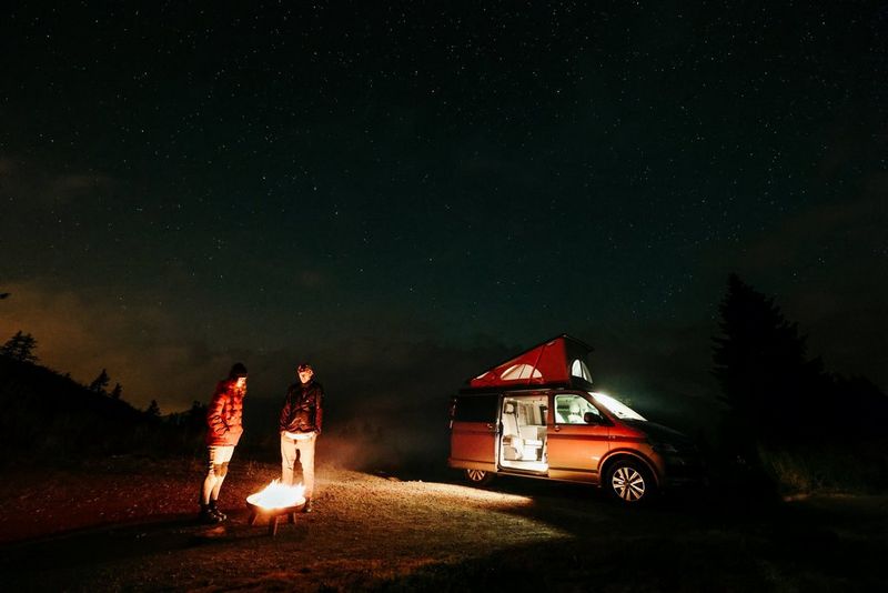 A low light image of two people around a firepit, lit by the glow of the fire, and in the background a campervan with interior lighting on.