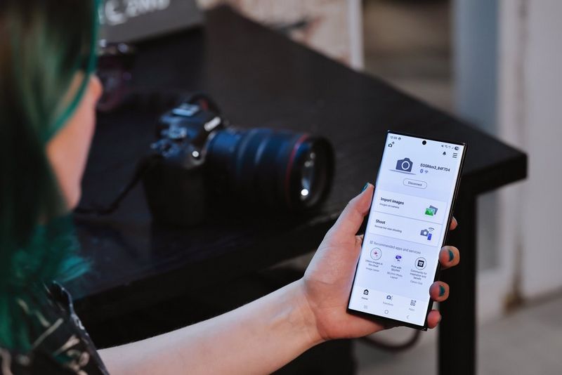 Un fotografo tiene in mano lo smartphone con l'app Canon Camera Connect aperta, mentre sul tavolo sullo sfondo è poggiata una fotocamera EOS R6 Mark III.