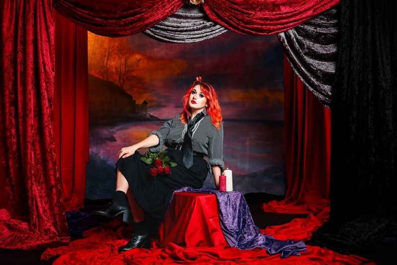 A richly colourful portrait photo of a woman with red hair sitting in a set draped with matching red and purple, taken by Corinne Cumming on a Canon EOS R6 Mark III with a Canon RF 24-105mm F2.8L IS USM Z lens.