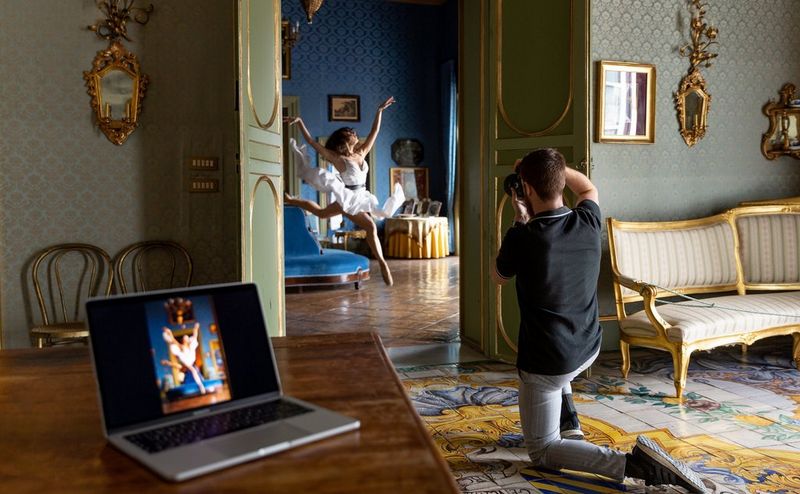 Javier Cortés zit geknield op één knie terwijl hij een balletdanseres filmt die een grote sprong maakt in een uitbundig versierde ruimte.