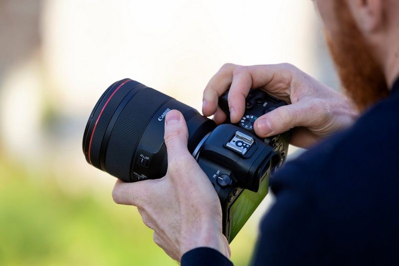 Een man houdt een Canon-camera met objectief vast terwijl hij met één hand het programmakeuzewiel aanpast.