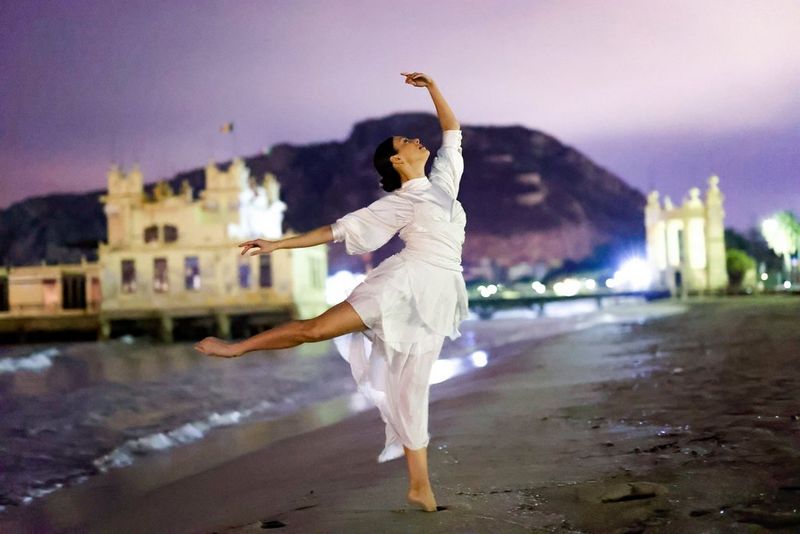 A ballerina dances on a moonlit beach with buildings lit up behind her.