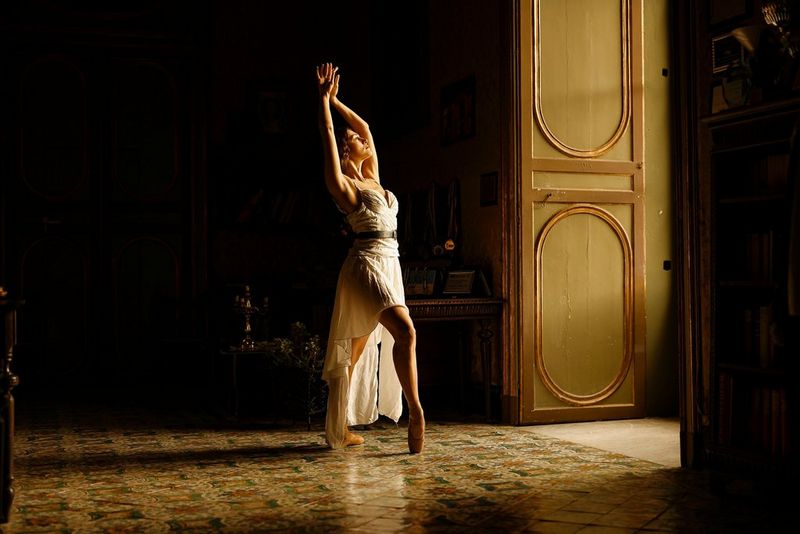 A ballerina poses in the light from an open door. The rest of the room is in shadow.