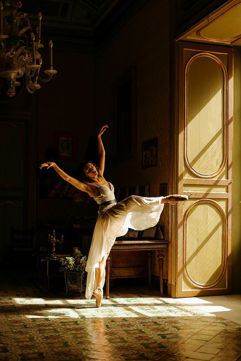 A photo taken on a Canon EOS R6 in available light showing a ballet dancer frozen in motion, standing en pointe on one foot, arms outstretched. She is lit by the light from a window to the side, with the room dark behind her.
