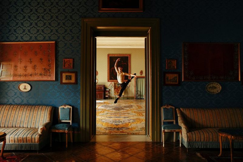 Een danser met een ontbloot bovenlijf springt omhoog voor een groot schilderij in een lijst, gezien door een open deur van een weelderig ingerichte ruimte.