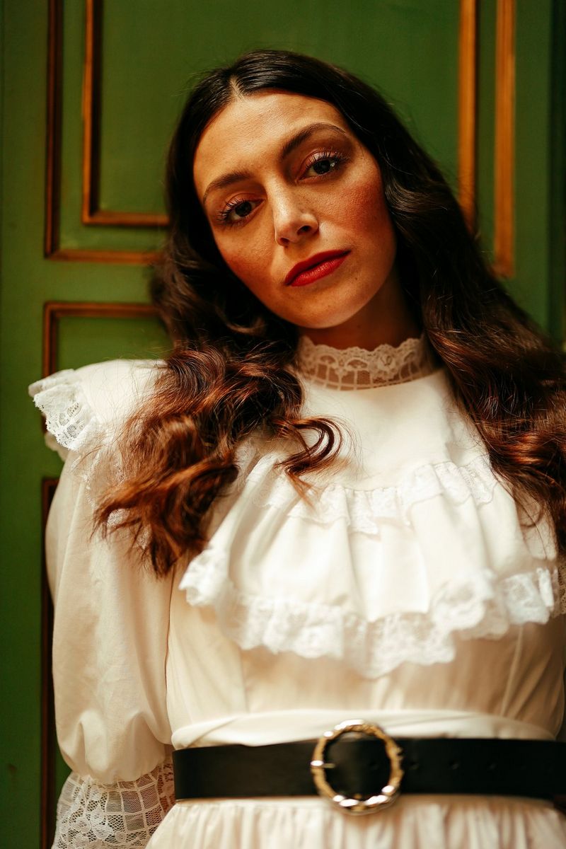 Una modella con lunghi capelli scuri e un abito bianco di pizzo con dei volant si appoggia a un muro verde e oro.