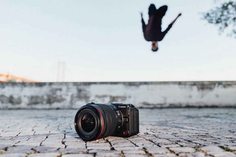 Камера Canon EOS R6 V с объективом Canon RF 20-50mm F4L IS USM PZ на вымощенной брусчаткой улице; на фоне можно увидеть, как паркурист выполняет сальто назад.