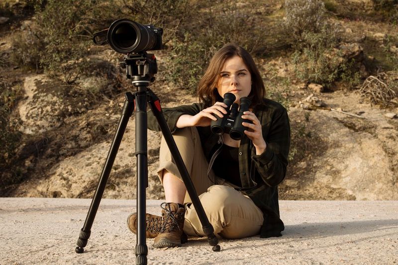 Dani Connor sits cross-legged on the ground with a pair of binoculars in her hands and a Canon EOS R7 on a tripod in front of her. 