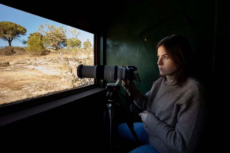 A woman sits in the shade of a shelter as she points her Canon camera, mounted on a tripod, out the window. 