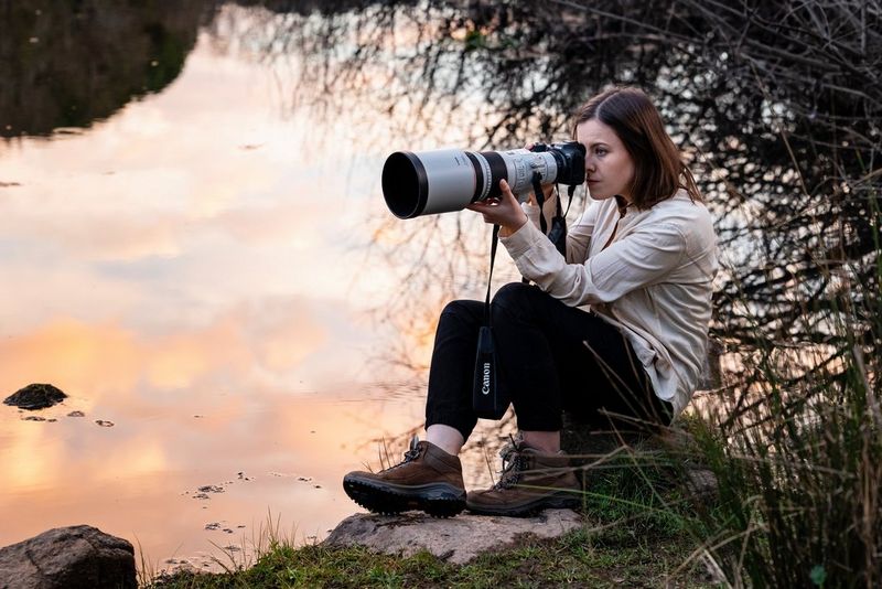 Dani Connor sits on a rock next to a lake taking photos with a Canon camera and white telephoto lens.
