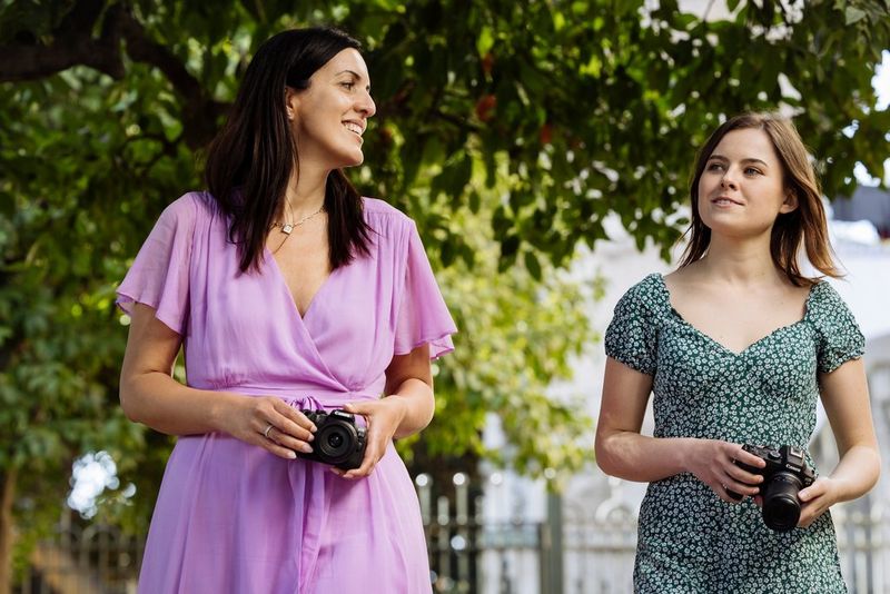 Two people, one wearing a light purple dress, the other a green dress with small white flowers, holding Canon cameras in a park.