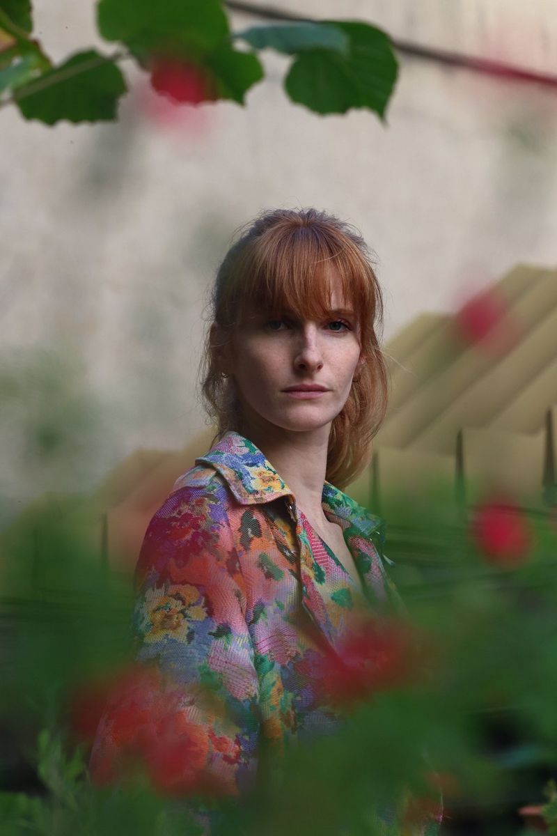 A woman in a colourful dress peeking through flowery bushes looks directly at the camera, shot on a Canon EOS R8. 