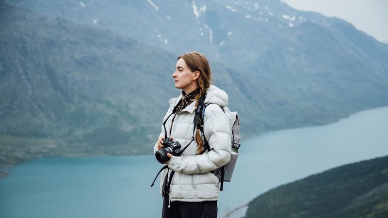 La photographe de voyage Frauke Hameister, vêtue chaudement, se tient au-dessus d'une crique norvégienne avec des montagnes en arrière-plan, un appareil photo Canon EOS R8 Mark II à la main.