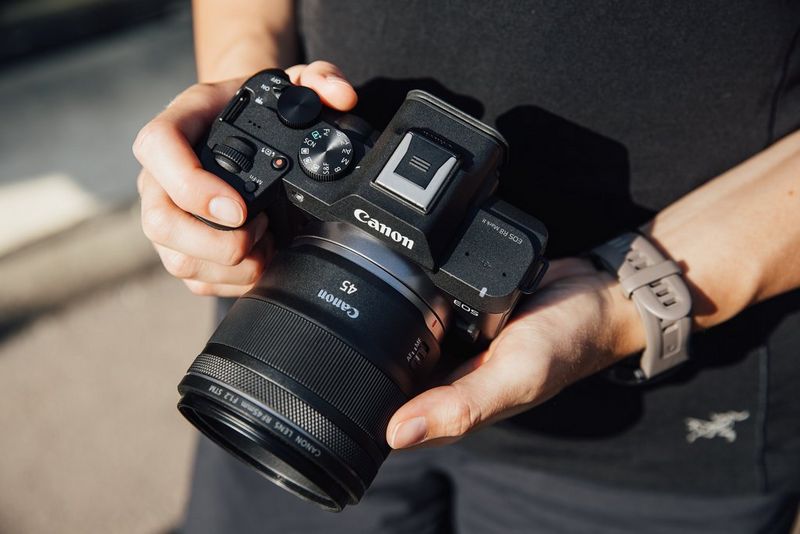 A woman holds a Canon EOS R8 Mark II with a Canon 45mm F1.2 STM attached, so that the dials and control switches on top of the camera are visible.