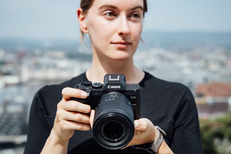 A woman holds a Canon EOS R8 Mark II full-frame camera, with an out-of-focus city view in the background.