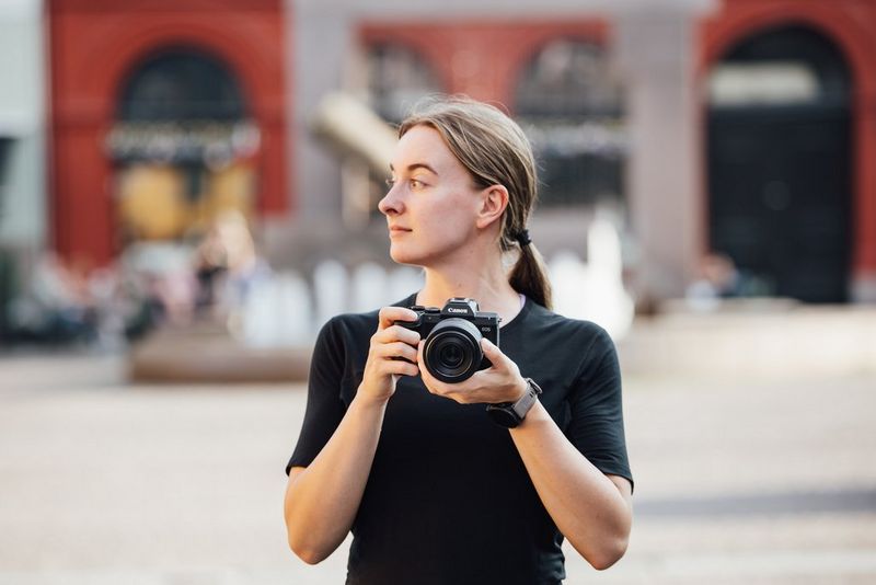 A woman holding a Canon EOS R8 Mark II, ready to take a photograph in a sunny urban setting.