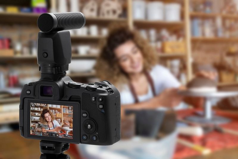 Een Canon EOS R8-camera met een externe microfoon op een statief filmt een persoon die iets op presenteert op een verhoging.