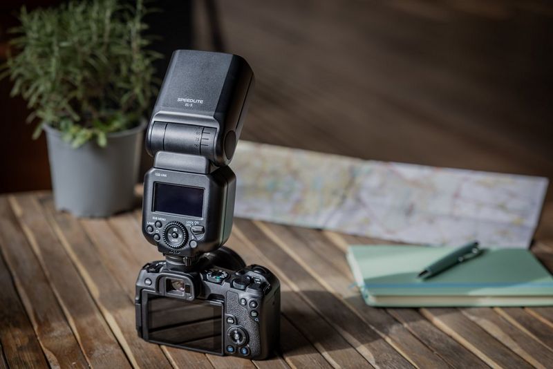 A Canon EOS R8 with a Speedlite flash fitted to its multi-function shoe sits on a table with a potted plant, map, notebook and pen next to it.