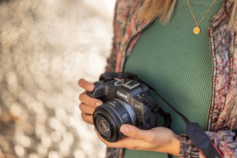 A person standing near a body of water holds a Canon EOS R8 in both hands.