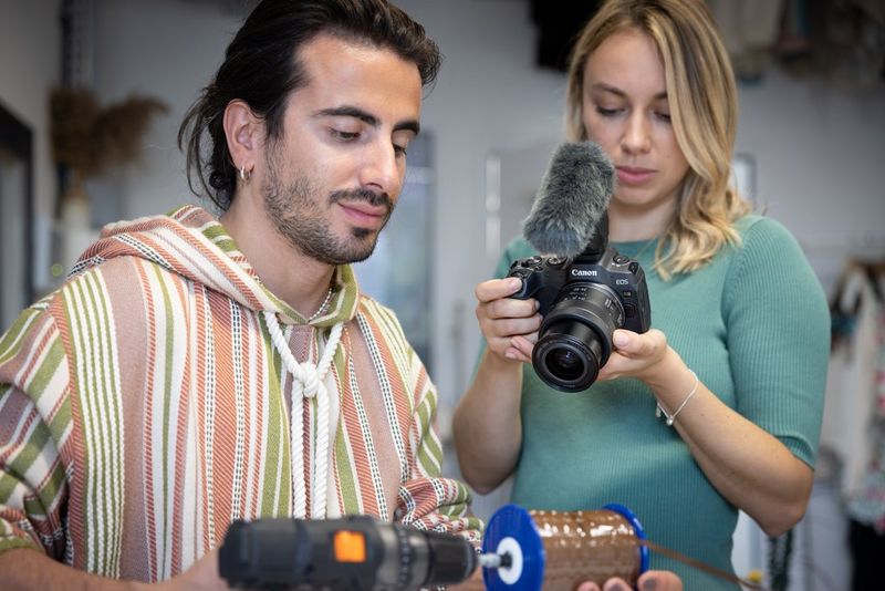 A woman uses a Canon EOS R8 with a mic attached to it to record a man using a power drill.