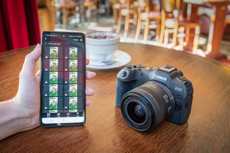 A hand holding a smartphone running the Canon Camera Connect app, with a Canon EOS R8 sitting alongside on a wooden table. 