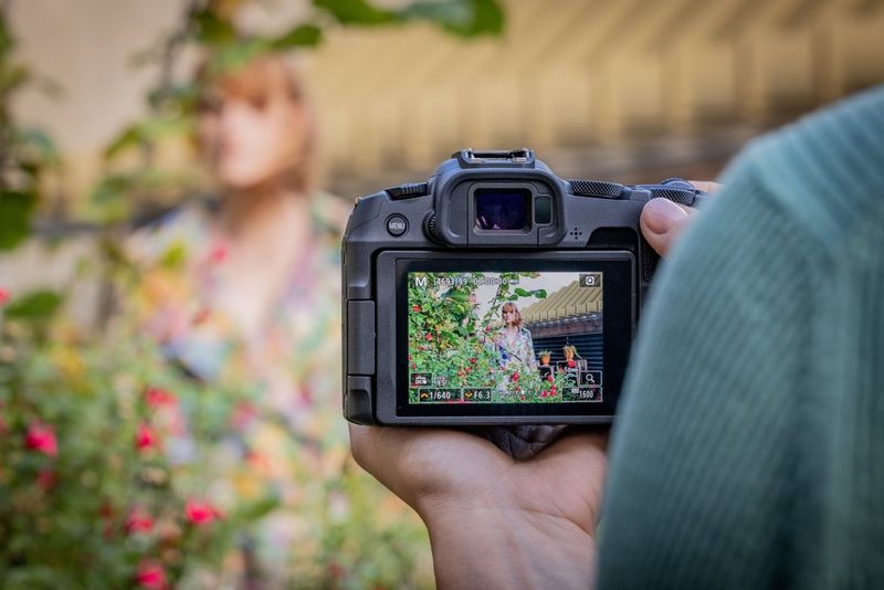 A view of the rear screen of a Canon EOS R8 being used to photograph a woman in a colourful dress standing next to a flowery bush.   